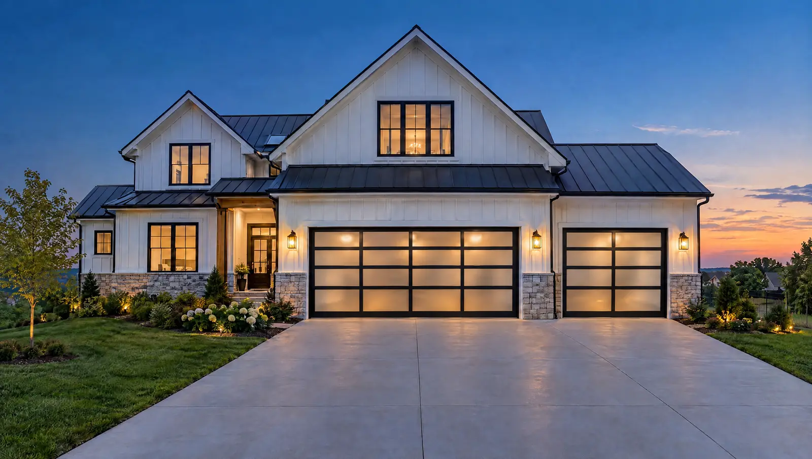 A white modern farmhouse at dusk with black full-view glass garage doors glowing from the lit garage